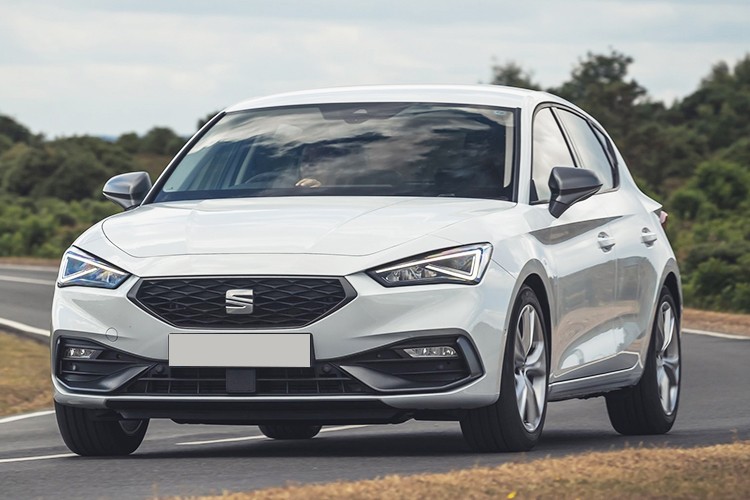 Front-side view of the SEAT Leon Hatchback in a crisp white finish, featuring sleek lines and dynamic LED headlights, captured on a winding road with greenery in the background.