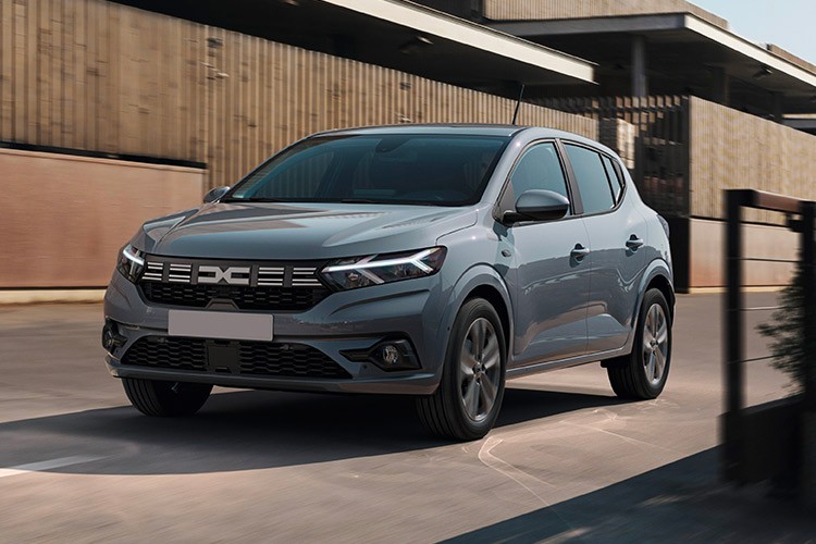 Side front view of the Dacia Sandero Hatchback in grey, highlighting its bold grille and modern headlights, set in an urban environment with contemporary architecture.